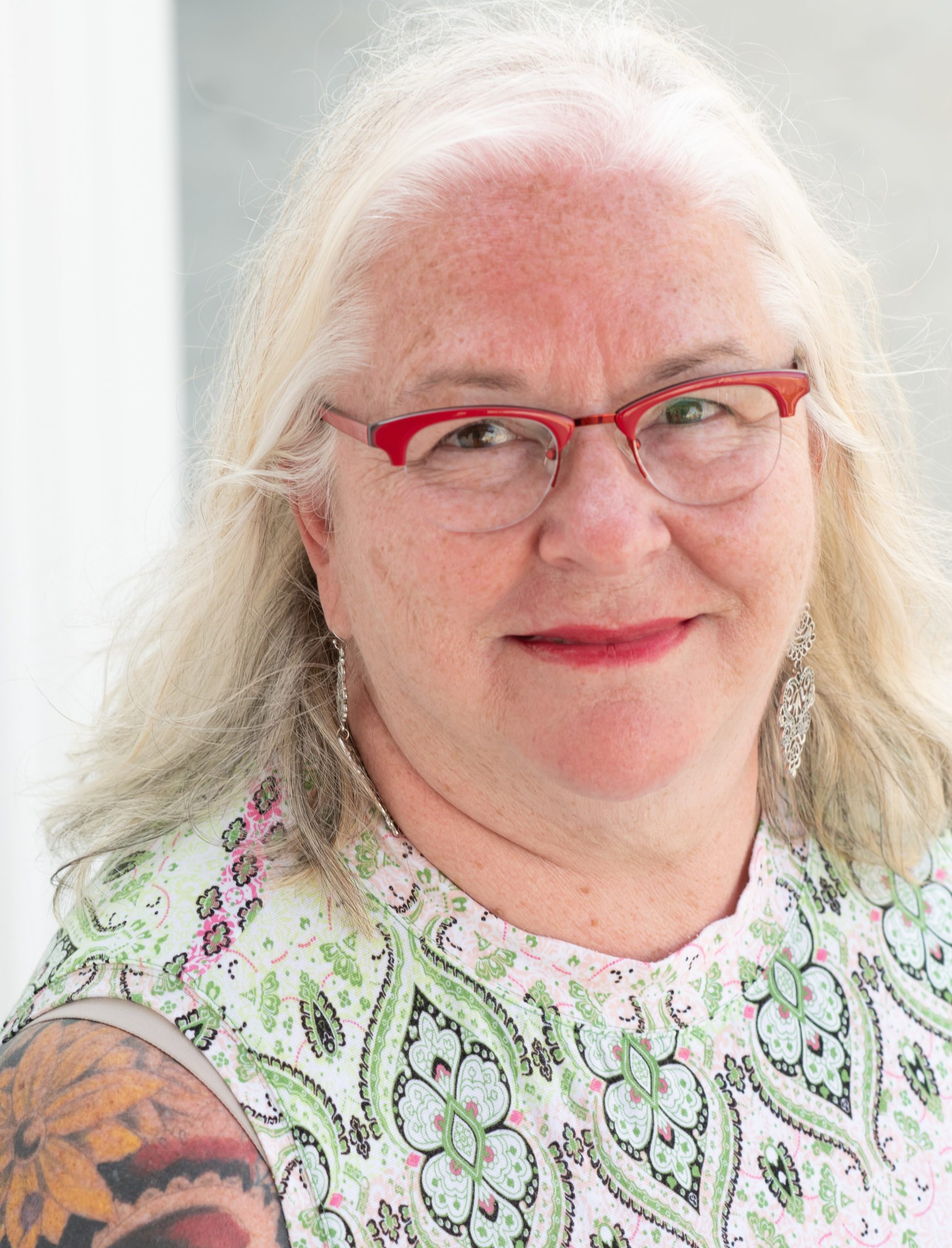 Deb Brown, a white woman with long white hair, poses in front of a light background, wearing red glasses, red lipstick, and a green and white printed sleeveless top