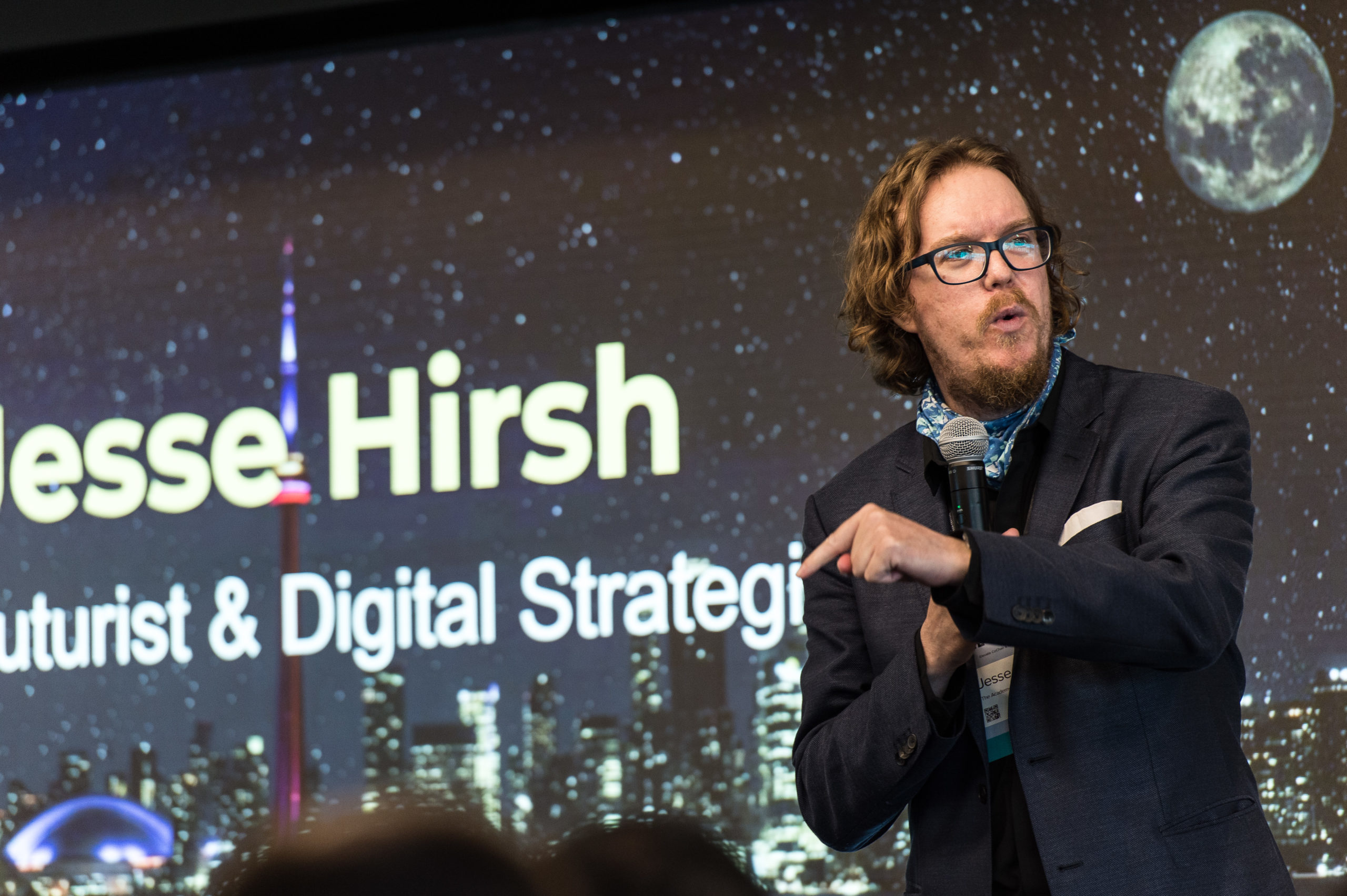 Jesse Hirsch speaks in front a screen bearing his name and the title "Futurist & Digital Strategist." Jesse is a white man with curly dark blonde hair and a beard and mustache. He is wearing glasses with thick black frames, a navy suit jacket over a black shirt and blue patterned scarf. He holds a microphone in his right hand and is gesturing with his left.