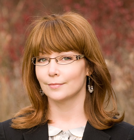 Margaret Doyle, a white woman with long brownish red hair and bangs, stands in front of a mottled brown background, wearing a black jacket, white top, and brown glasses
