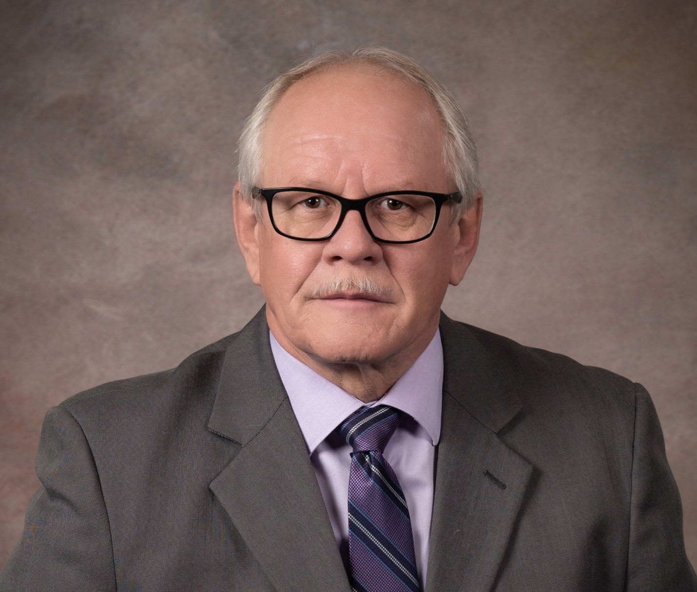 Ron McCullough, a white man with white hair on the sides and a white moustache, sits in front of a mottled brown background, wearing black glasses, a grey suit, a purple shirt, and a patterned purple tie